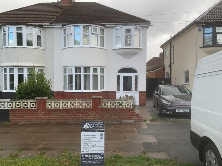 Semi-detached house rendered in smooth white with a red brick plinth and white bay windows, AJC and Son Builders sign board on the front path